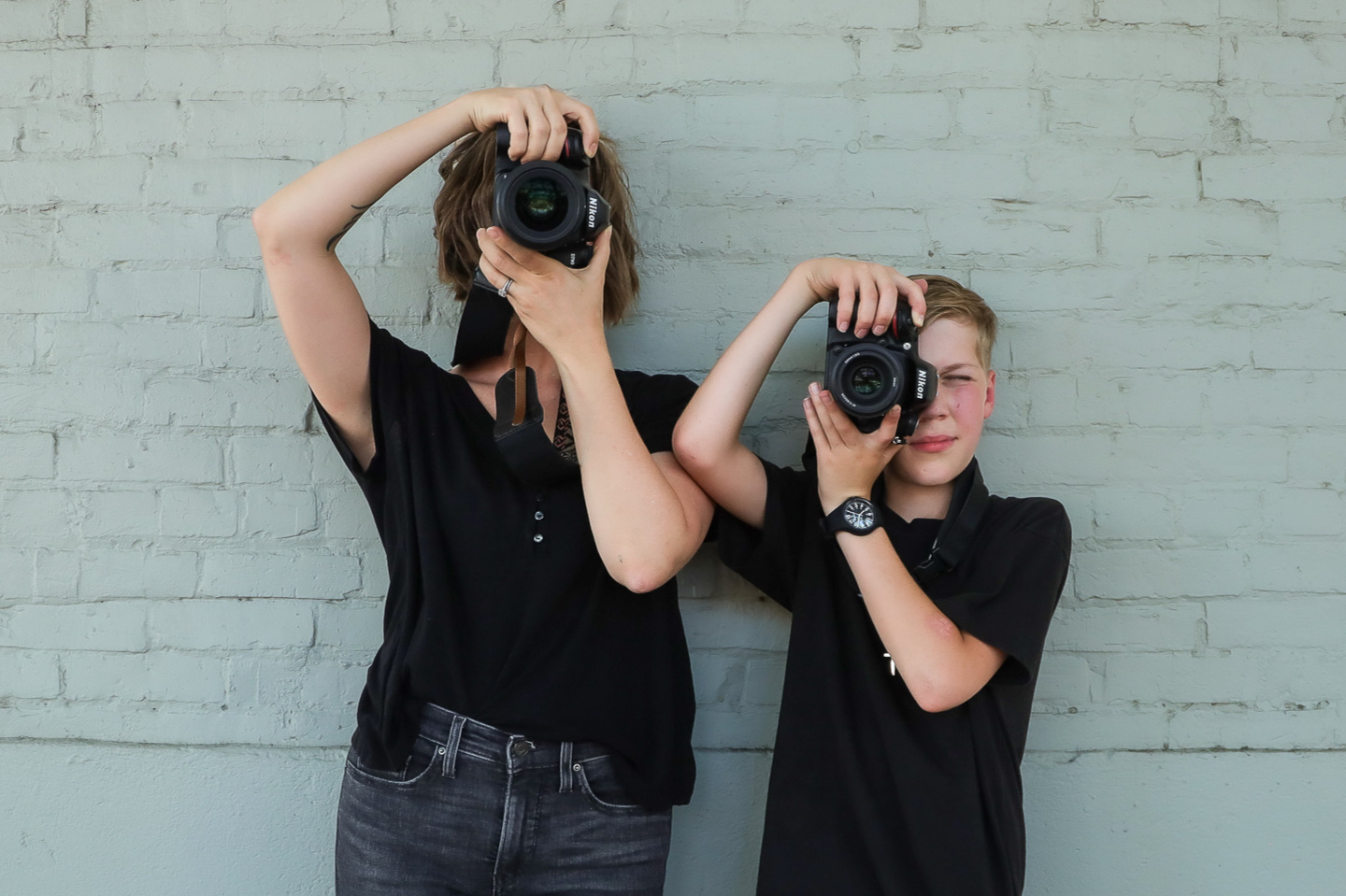 image of professional mother son photographer team against a cream brick wall; they are holding their cameras to their faces.