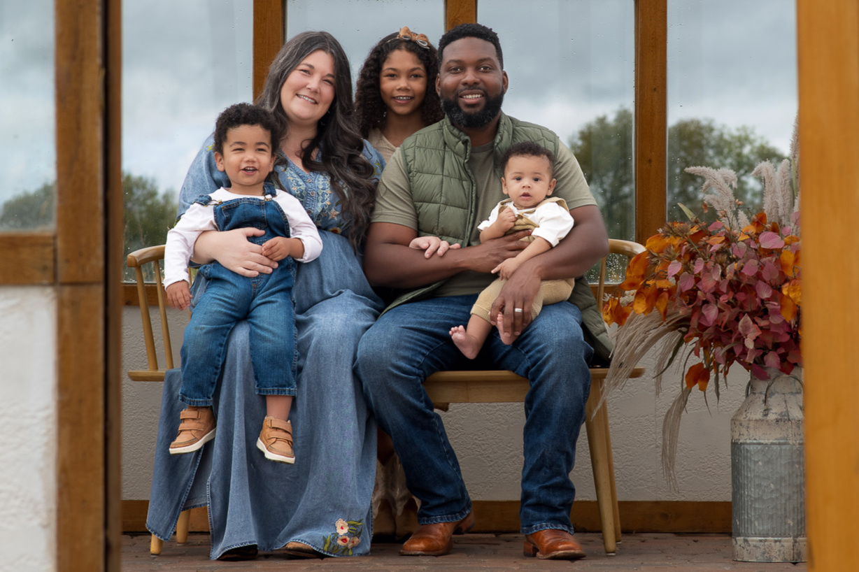 Mother and father with three children cuddled on a bench together in an outdoor greenhouse with fall themed wood and florals at a lifestyle family photography session in Waxahachie, TX, southern DFW.